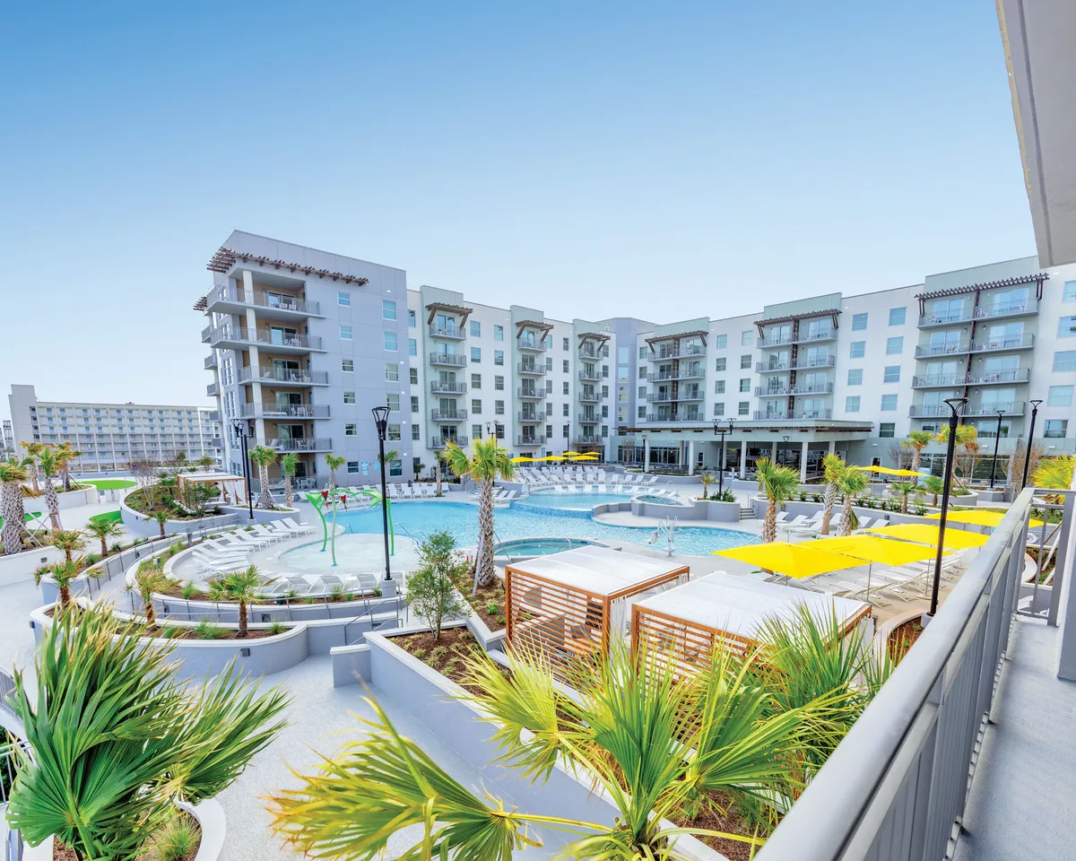 A lively resort pool scene featuring a large, sparkling swimming pool surrounded by palm trees and modern apartment buildings. Yellow umbrellas and poolside cabanas offer shade, while guests are seen sunbathing, socializing, and relaxing in the cheerful vacation atmosphere.