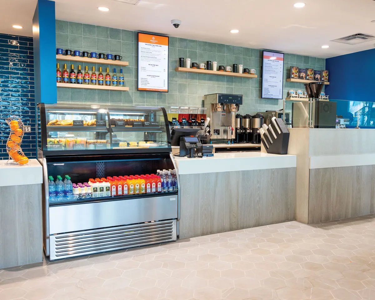This image features a well-lit interior area with a display case full of fresh pastries, a beverage cooler, and coffee machines. There are menu boards overhead and decorative tiles that give the space a welcoming vibe. People are seen grabbing coffee, socializing, and working on laptops.