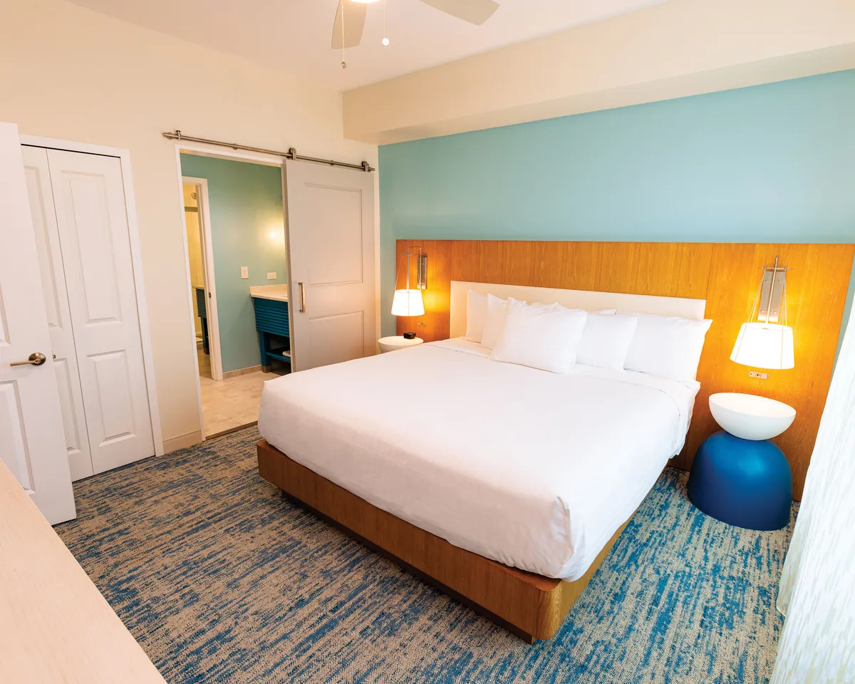 The image features a beautifully appointed hotel bedroom with a plush bed, soft lighting from modern fixtures, and a cozy nightstand—creating a calm and inviting atmosphere perfect for unwinding after a day at the beach.