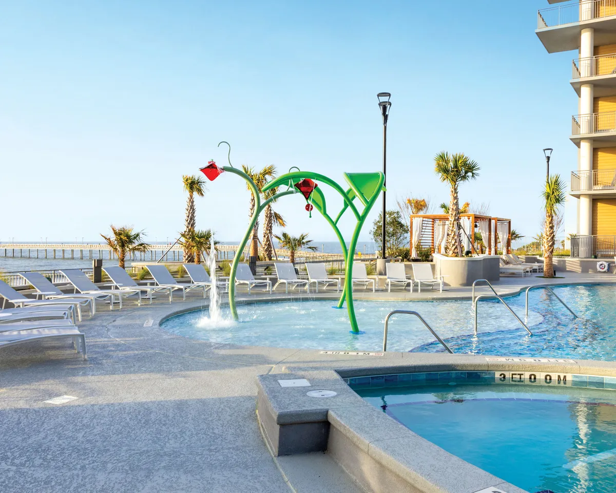 A vibrant family-friendly swimming pool area featuring a water play structure for kids, surrounded by lounge chairs and palm trees with a beautiful view of the ocean in the distance. Children and families are engaged in water play and sunbathing, creating an atmosphere of joy and relaxation.