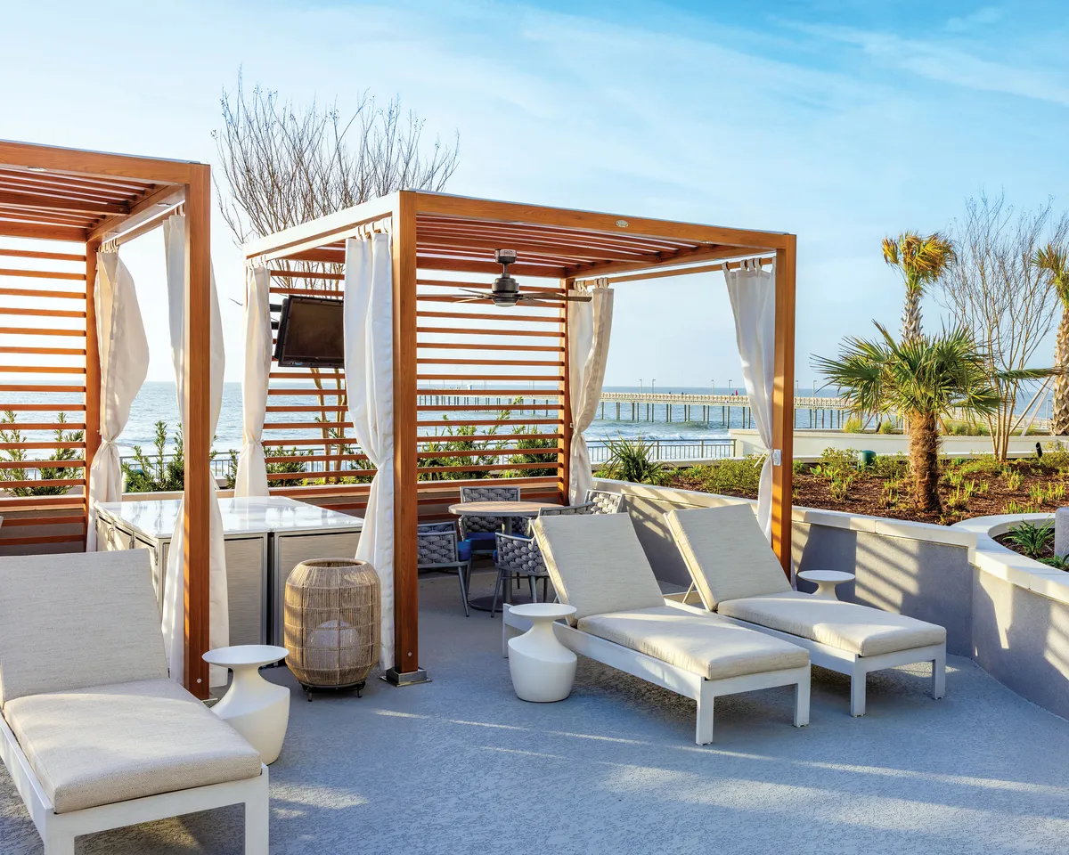 A tranquil balcony scene featuring wooden cabanas and lounge chairs set against a sweeping view of the Atlantic Ocean. Palm trees gently frame the balcony, and the horizon hints at the potential for both sunrise coffees and sunset relaxation. The space appears inviting for both intimate gatherings and quiet moments, underscoring the idea of an extended living area.