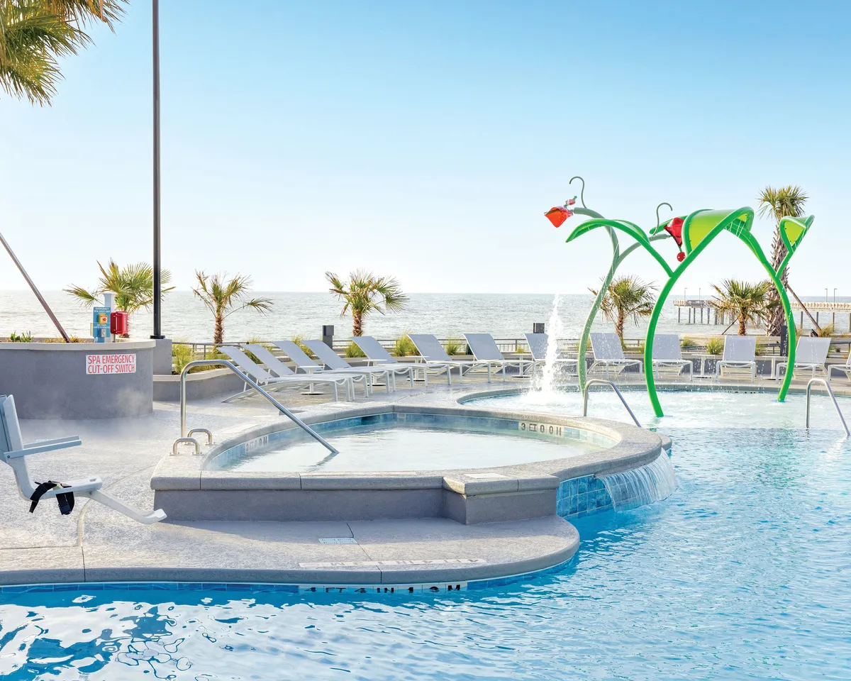 A lively exterior setting where a pool area with colorful play structures is visible below, bordered by lounge chairs and an ocean view in the background. The atmosphere is playful and serene, suggesting both family fun and relaxation. The placement of the balcony (from which the scene might be observed) is implied but not shown directly, connecting to the idea of watching activities below.