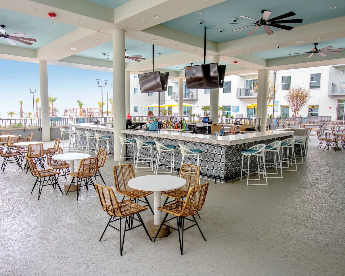 An airy poolside bar area with comfortable seating, ceiling fans, and large windows, set up for socializing and relaxation. Guests are enjoying drinks and friendly conversation as the sun sets, creating a cheerful and inviting atmosphere.
