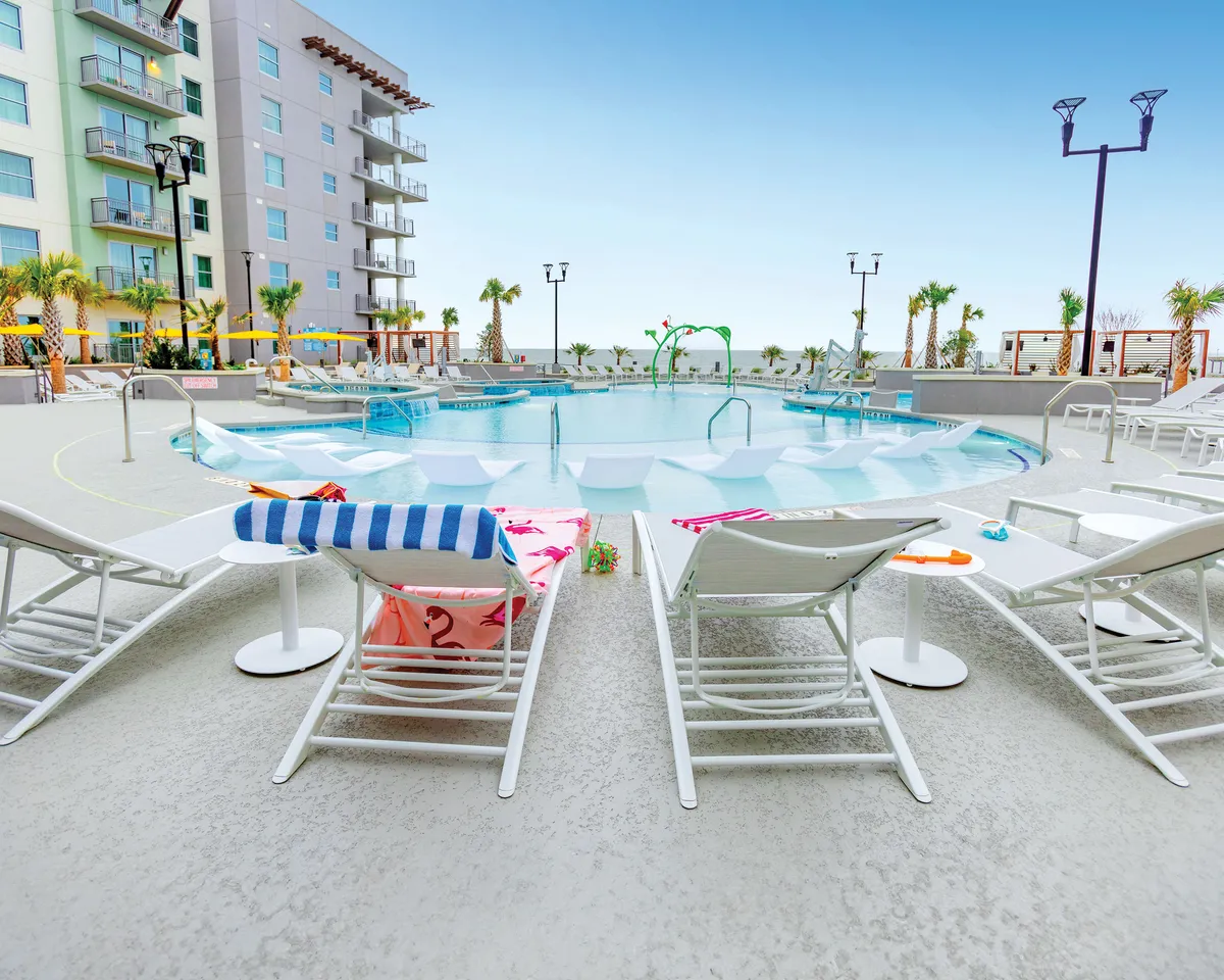 A sun-soaked pool area with comfortable lounge chairs draped in colorful towels, surrounded by palm trees. Children play in the water while others enjoy swimming, sunbathing, and socializing in a carefree environment.