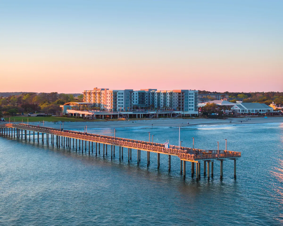 A scenic view of a pier stretching over the water, with nearby buildings and an expansive sky. People can be seen strolling, fishing, and enjoying the relaxed coastal ambiance—ideal for sunset walks and sightseeing by the shore.