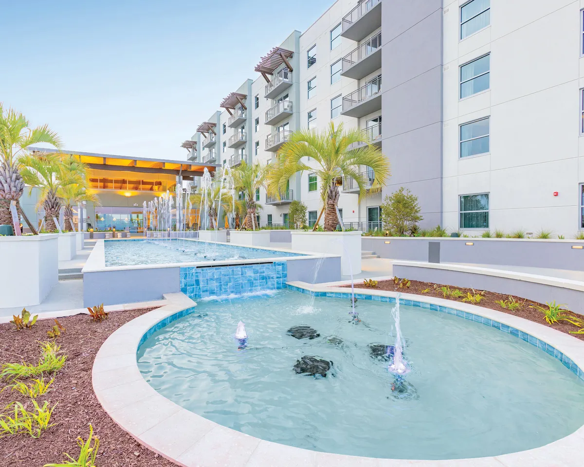 This image captures the exterior of the property, featuring elegant water features, swaying palm trees, and striking modern architecture. The tranquil setting highlights sunset views by the pool and a vibrant, inviting courtyard where guests can relax or socialize. The scene evokes a sense of escape and romance, with natural beauty and luxurious resort amenities on display.